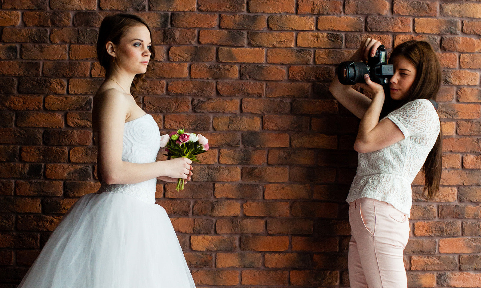 Wedding portrait photoshoot with bride holding flowers and photographer capturing images in front of a brick wall.