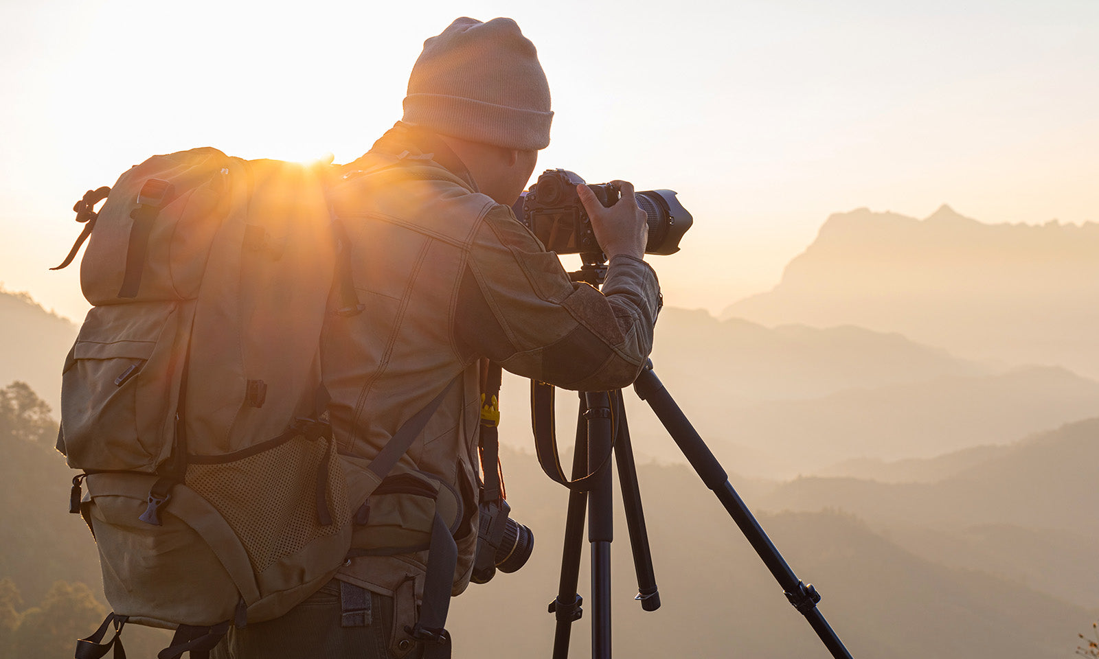 Travel photographer taking sunrise mountain photos with a DSLR camera, tripod, and backpack in a scenic outdoor location.